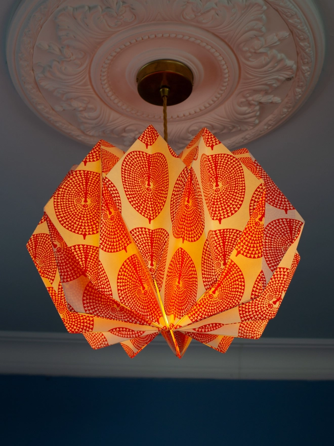 A vintage-inspired paper lampshade with a bold geometric pattern adds a touch of retro flair to a room with a blue wall and ornate ceiling. The lampshade is made from folded paper, creating a unique and eye-catching design.