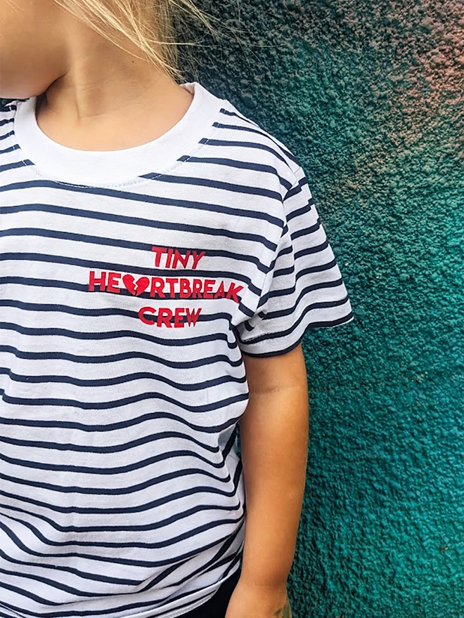 Child wearing a navy and white stripe t-shirt with red Tiny Heartbreak Crew slogan.