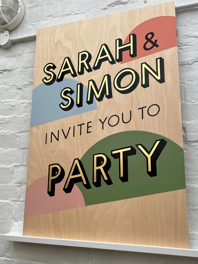 Handpainted invite to party sign with gold leaf letters outlined in black, with coloured semicircles in the background, against a white brick wall. 