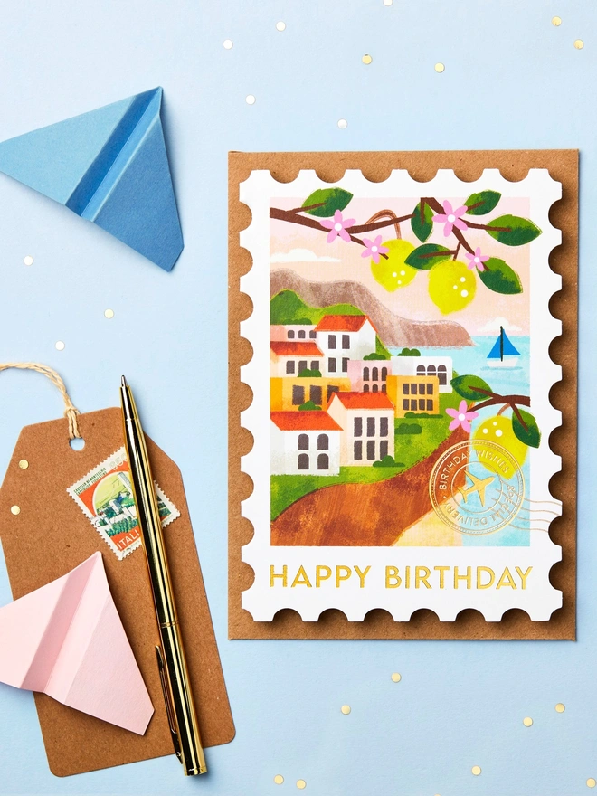 Stamp-shaped greeting card featuring an illustration of the Italian Riviera. The card is on a blue background with paper aeroplanes and stationery around it.