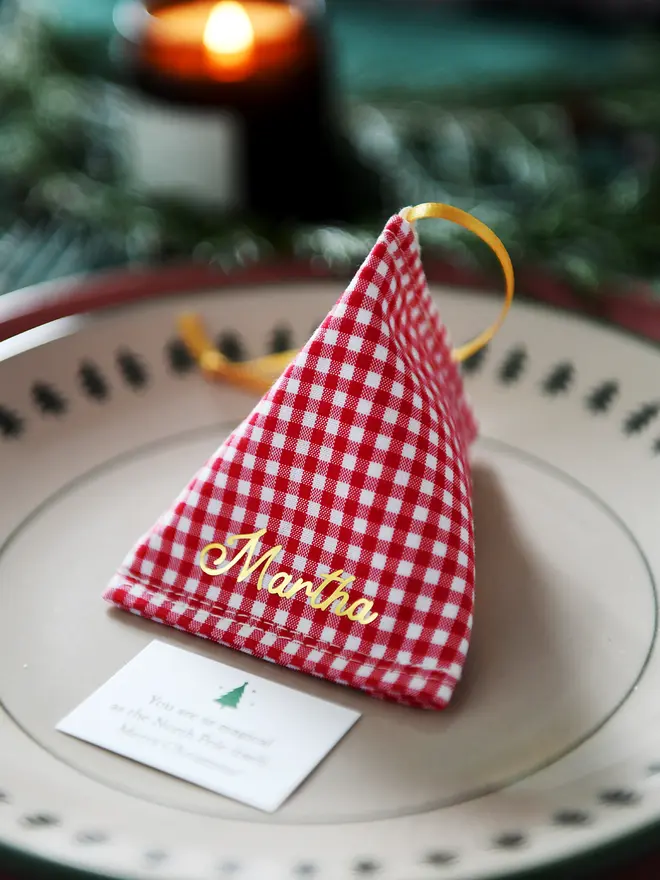 A red gingham fabric fortune cookie with gold personalised name “Martha” sits on a Christmas dinner plate, ready as a handmade festive place setting with a hidden Christmas wish card beneath.