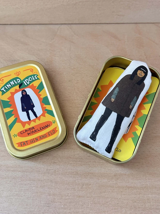 Mini fabric doll featuring an illustration of Claudia Winkleman in her Traitors cape. The doll is beside a gift tin.