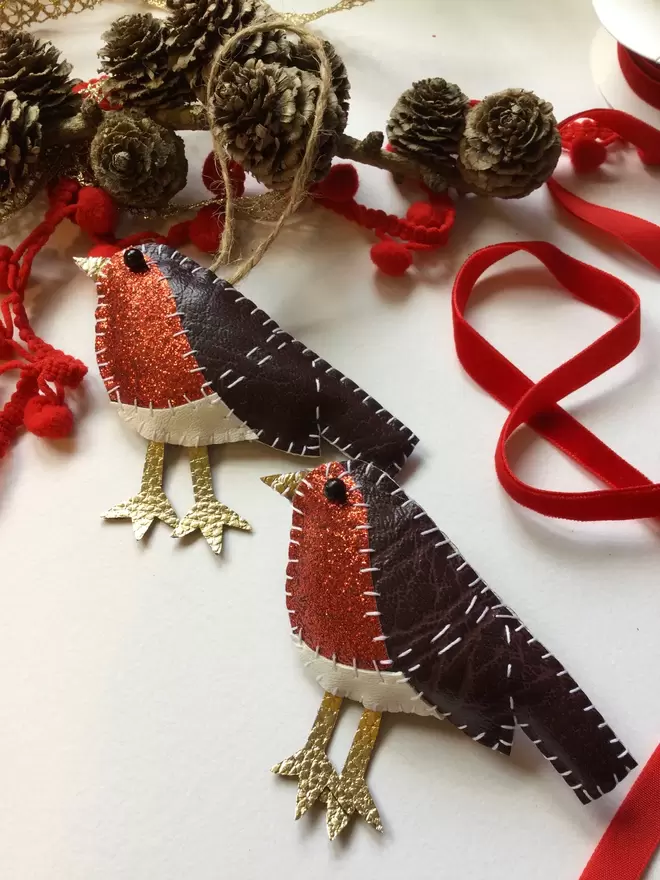 A leatherette robin Christmas tree decoration and brooch, hand stitched, with red glittery breast, sits amongst pine cones, red ribbon, and pom pom trim,