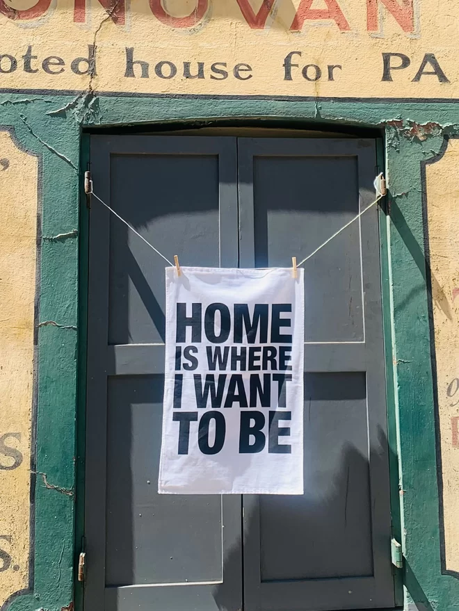 London Drying Home Is Where I Want to Be black screen printed text on white tea towel hanging in front of a disused shop front