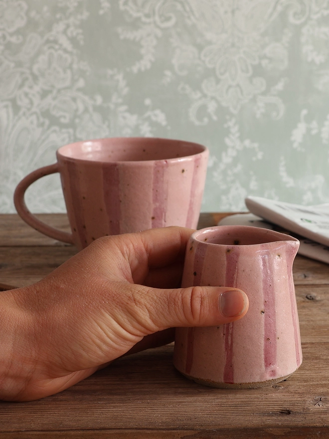 Hand holding a Handmade Ceramic Stoneware Small Jug in Pink Stripe which is sat on a wooden surface with a mug in the same style and a newspaper behind it. 
