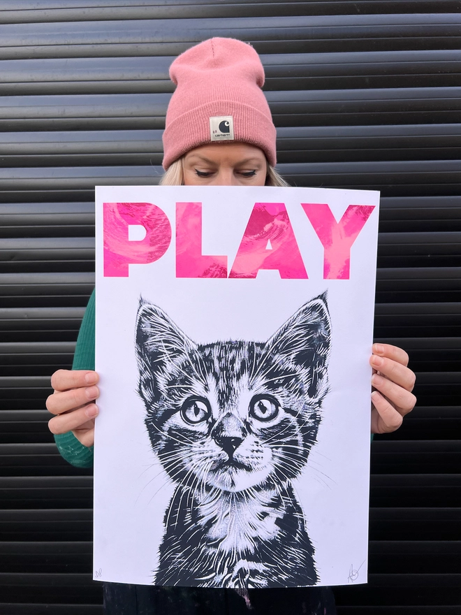 woman artist in pink beanie holding original artwork of a cute kitten, with the word play mono printed above, screen print and mono prit, cute art, art for kids room