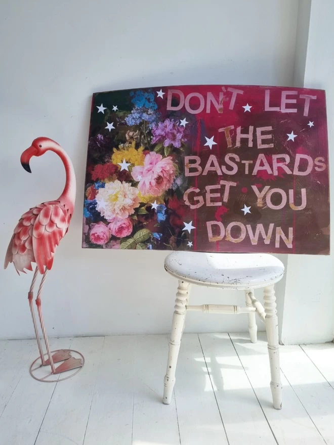 Art panel made from an upcycled table top and decorated using decoupage florals in pinks and browns. The text reads ‘don’t let the bastards get you down’ in the hot pink undercoat. Decorated with stars.