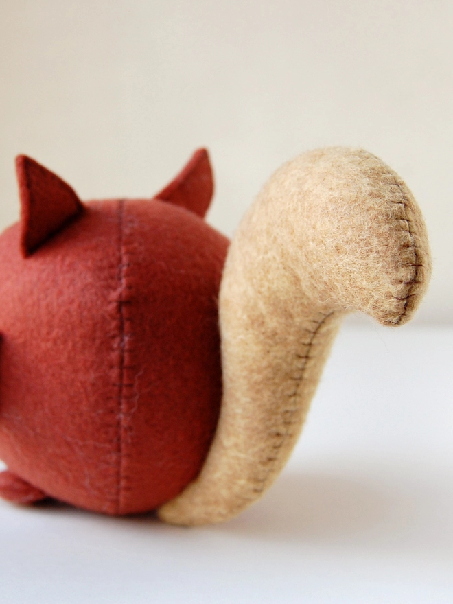 A handmade felt squirrel toy faces backwards with a large felt tail, on a white wooden desk.