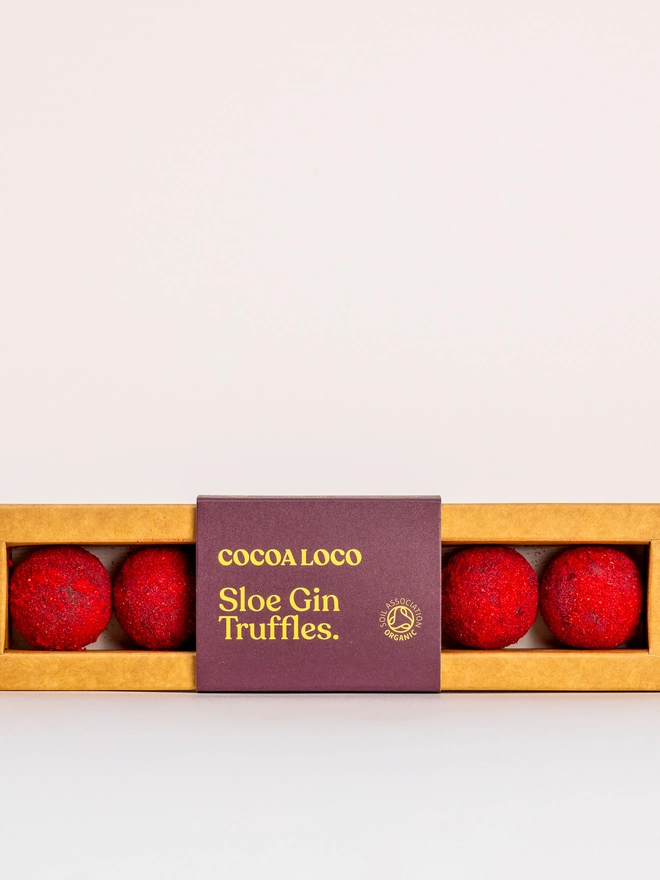 Milk Chocolate Sloe Gin Truffles, a box of truffles sitting on a white surface, packaged together. They are wrapped by a colourful logo. 