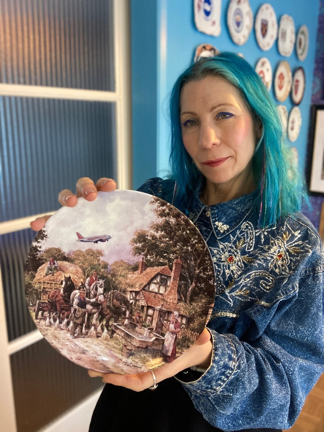 Lucy is holding the decorative 8” plate featuring a chocolate box-twee cottage and a hay cart pulled by shirehorses. An easyJet plane soars off in the distance, causing a jarring contrast to the 17th century rural scene. 