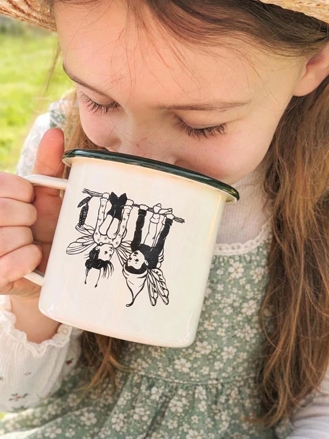 girl drinking from an enamel mug cream in colour with a green rim etched with two pixie pals
