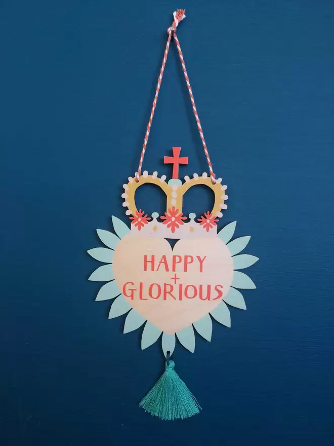 'Happy + Glorious' is hand printed on a colourful wooden heart with party crown and tassel. 