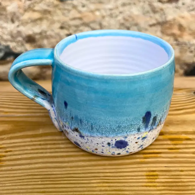 Shoreline Large Round Mug, a colourful mug on a wooden surface against a brick backdrop. 