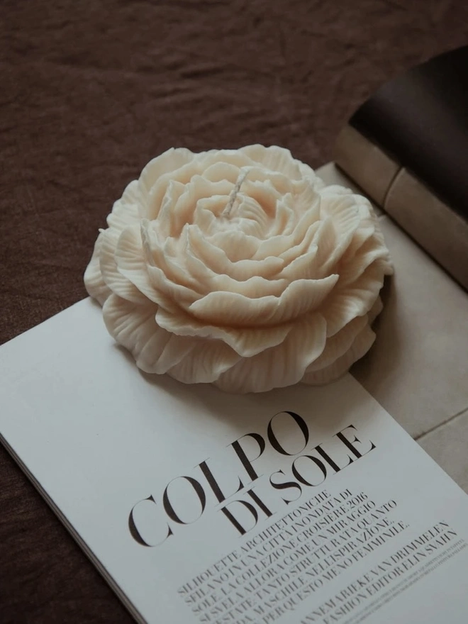 Large White Peony Candle. A cream peony-shaped candle with layers of finely detailed petals, resting on an open magazine beside a wine glass on a brown linen cloth. The realistic floral design makes an elegant decorative piece for a coffee table or bedside.