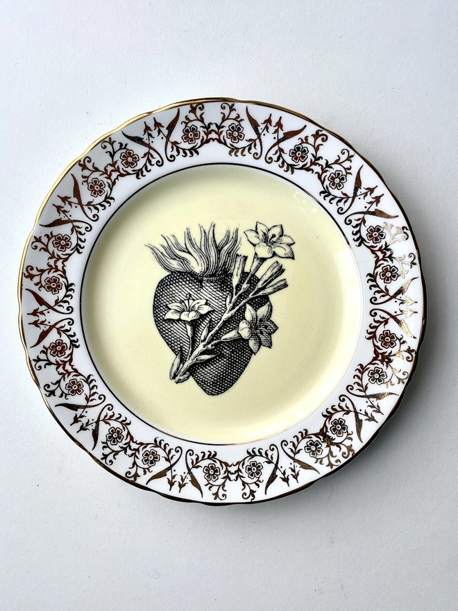 a vintage plate with a white and gold ornate border, the centre is lemon yellow and printed on that is a black illustration of a sacred heart