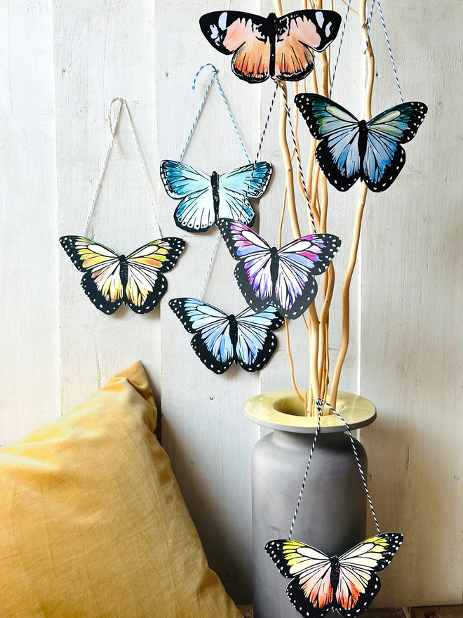Hand Painted Butterfly Colourful Linocut Hanging Decoration multiple butterflies hanging on a display of sticks in a vase for context of how they can be hung in your home and two on a wall with yellow cushion in front
