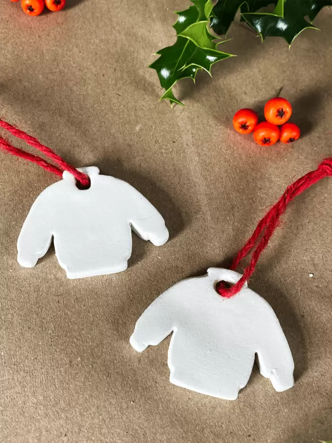 Two white clay christmas jumper tags with red twine on a brown parcel paper background with some holly and berries in the top half of the image.