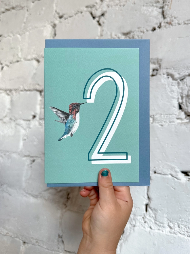 a pale teal card with a big white number 2 and an illustration of a hummingbird with a blue envelope