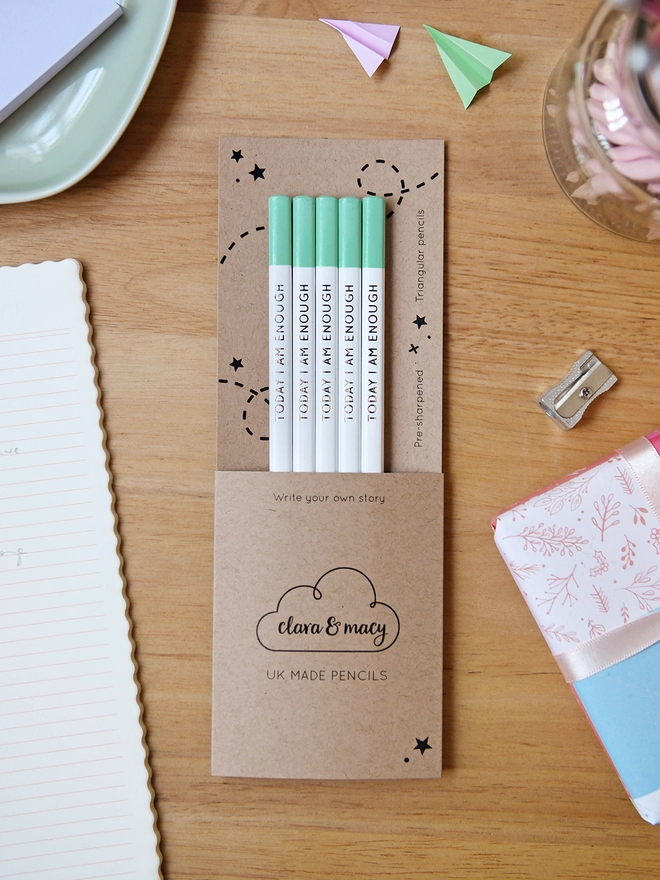 Five white pencils with green tips and the words Today I Am Enough written along the side, sit within cardboard packaging on a wooden desk with stationery items.