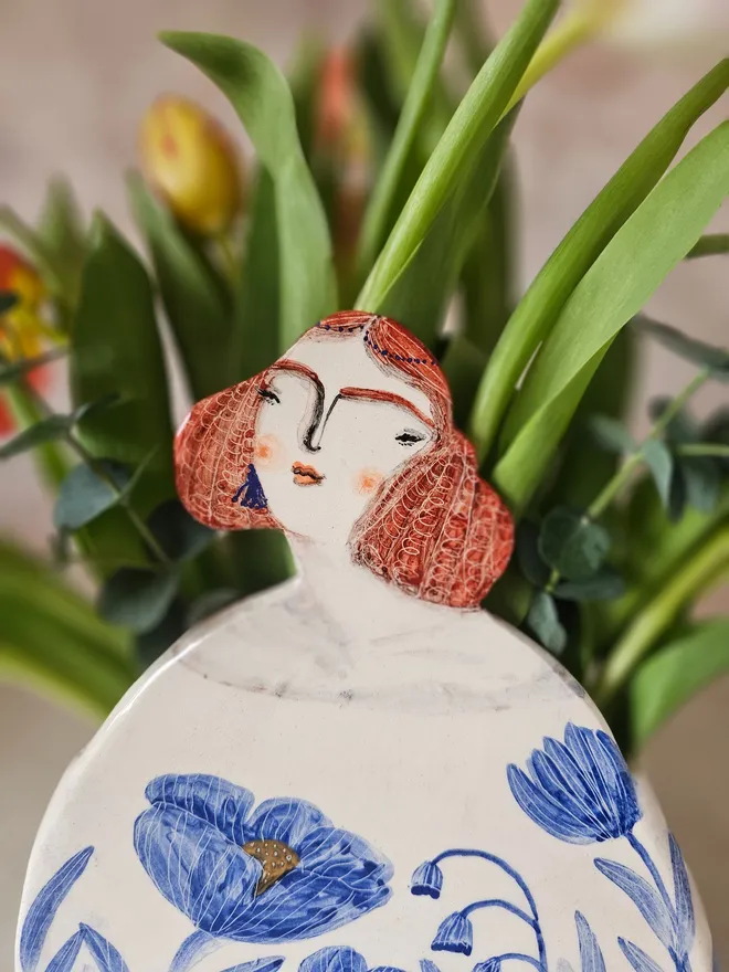 a large vase shaped like a lady with red hair looking up with blue flowers painted on her dress, front view close up