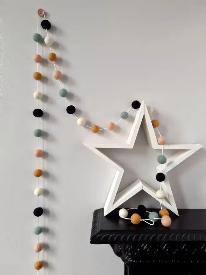 A white wooden star on a black mantelpiece, with a felt ball bunting hanging above in soft, Halloween shades.