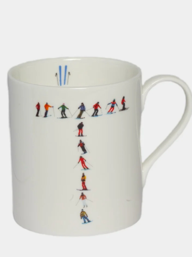 A simple white mug illustrated with a ’T’ made of small, multicoloured skiers. The mug is set against a plain, white background.