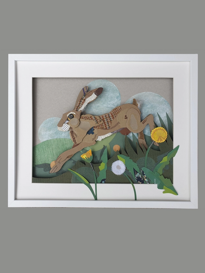 Hare leaping through long grass and dandelions, created using multiple layers of corrugated card, decorated with salvaged fabric and paper. Including buttons and feathers. Finished off with machine and hand embroidery.  