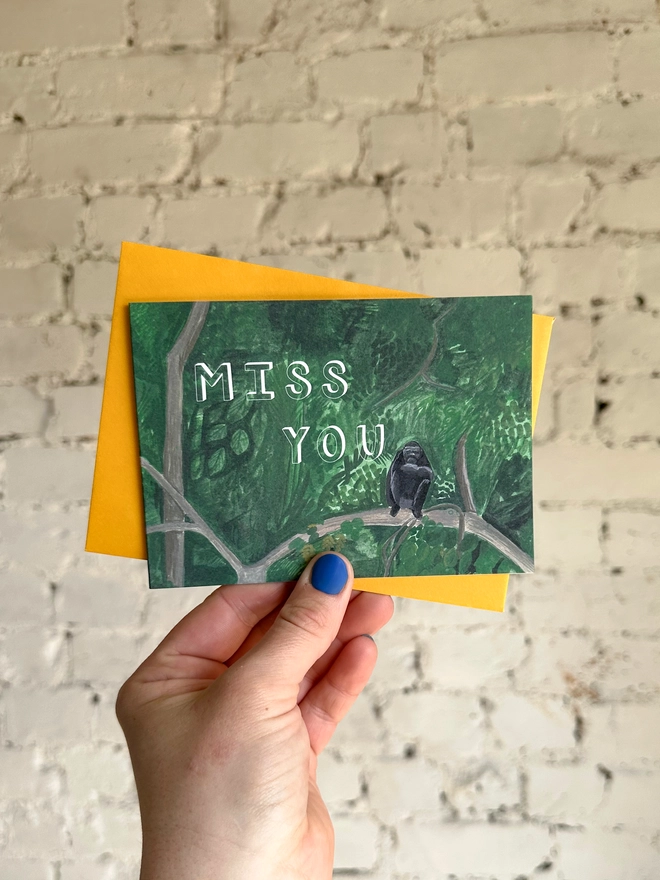 a greetings card featuring an illustration of a chimp sitting in a jungle landscape with the text “miss you” with a deep yellow envelope