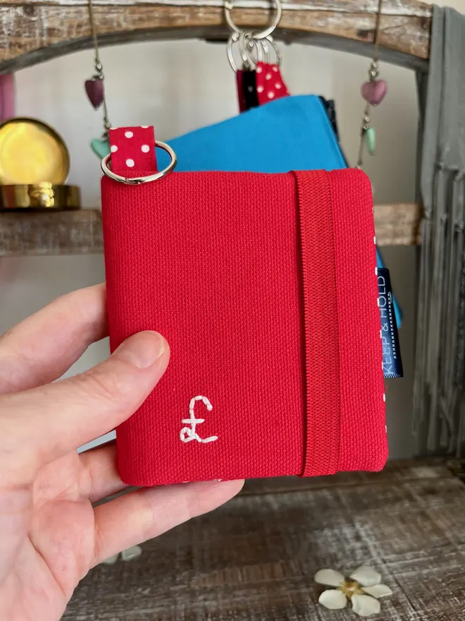 Hand holding a small red card wallet with keyring and red elastic closure. A £ sign embroidered on the front in white thread.