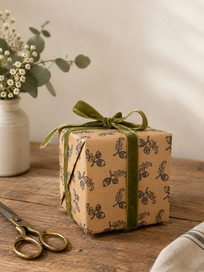 A PHOTO OF A SQUARE PRESENT WRAPPED IN KRAFT PAPER STAMPED WITH A SWEET ACORN PATTERN TIED WITH A GREEN VELVET RIBBON
