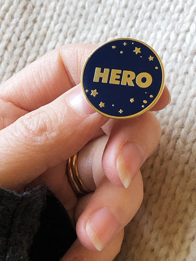 A navy blue and gold enamel pin badge with a starry design and the word "Hero" is being held.