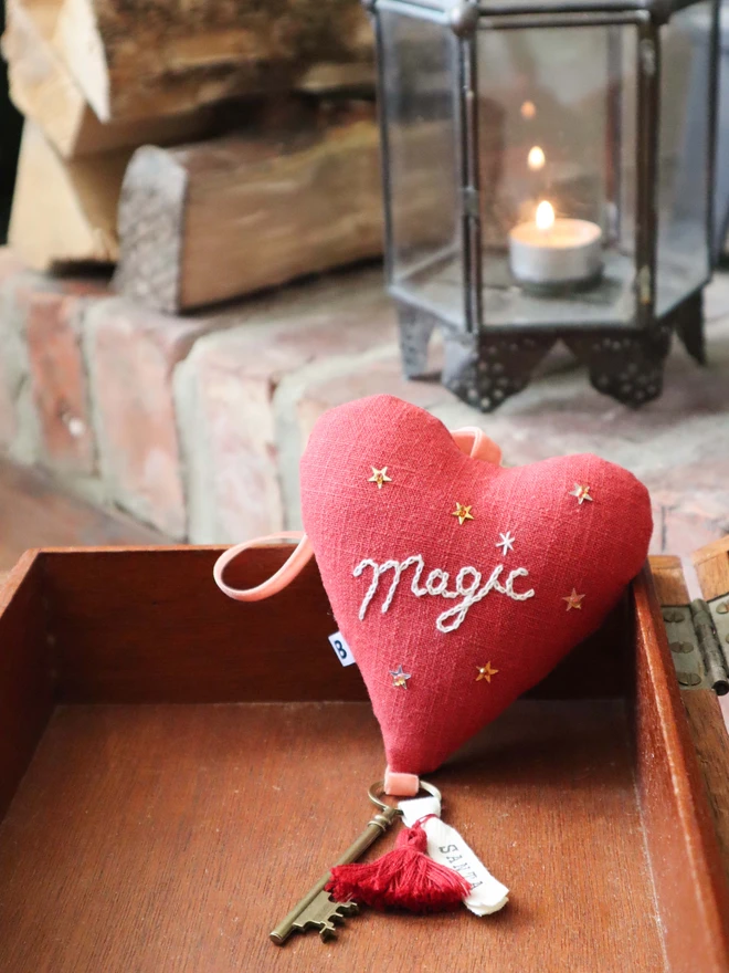 Linen heart ornament with Magic embroidery, hanging rustic key with Santa's name tag and red tassles, surrounded by Christmas presents and elf doll. 