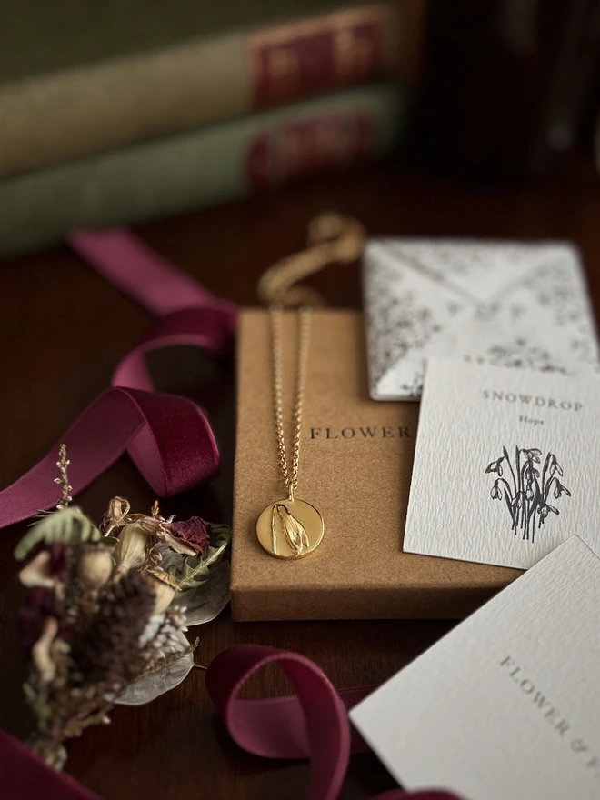 Gold snowdrop coin belcher necklace representing hope draped over flower & fable packaging