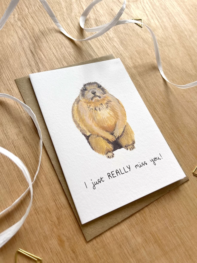 a greetings card with a white background featuring an illustration of a sad looking prairie dog with the phrase “I just really miss you”
