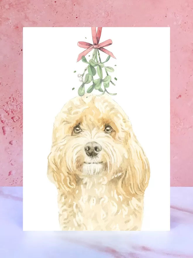 A Christmas card featuring a hand painted design of a Apricot Cavapoo, stood upright on a marble surface.