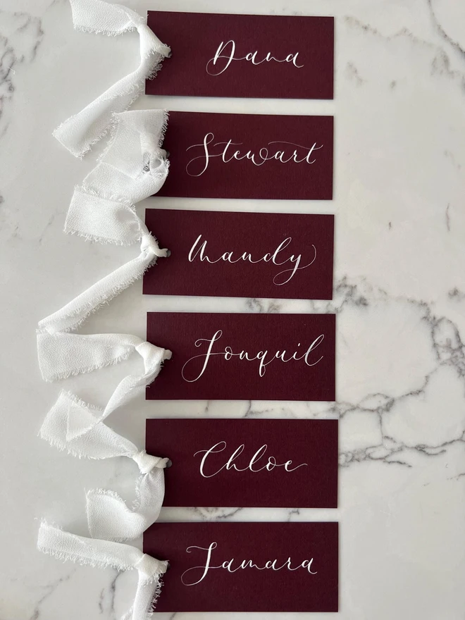 Personalised Calligraphy Wedding Burgundy Place Cards, burgundy place cards set together on a wooden table with white ribbons. 