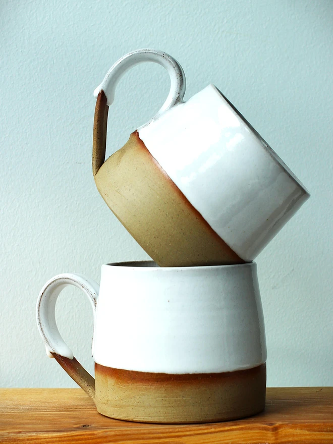 Handmade ceramic mug with a glossy white upper glaze and a natural toasted clay base, against a soft green wall background.