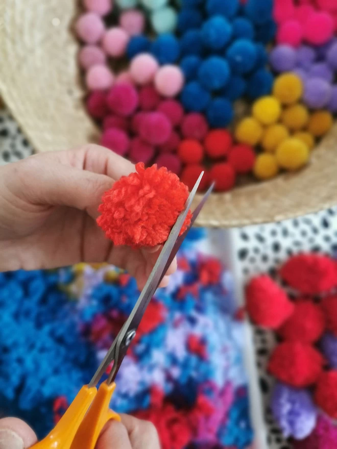 An overhead image of a hand trimming a homemade pom pom with orange handled scissors.. Behind the image is a woven basket bursting with multi coloured pom poms.