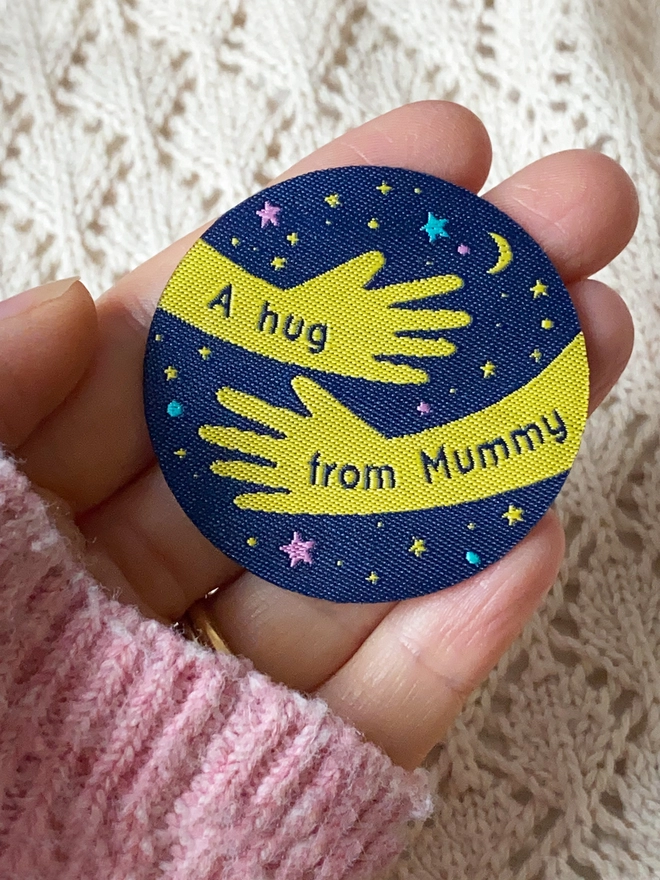 A navy blue and yellow woven patch is being held on an open hand. The design is two arms in a hug shape with the words "A hug from Mummy" along them. 