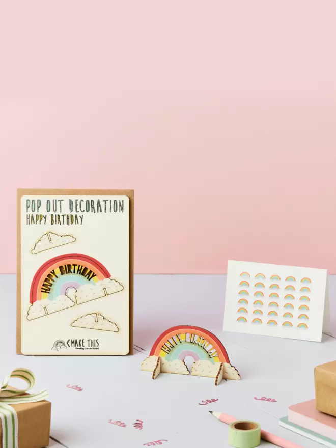 3D rainbow birthday decoration with laser-cut Happy Birthday text and rainbow pattern birthday card and brown kraft envelope on top of a grey desk in front of a pink coloured background