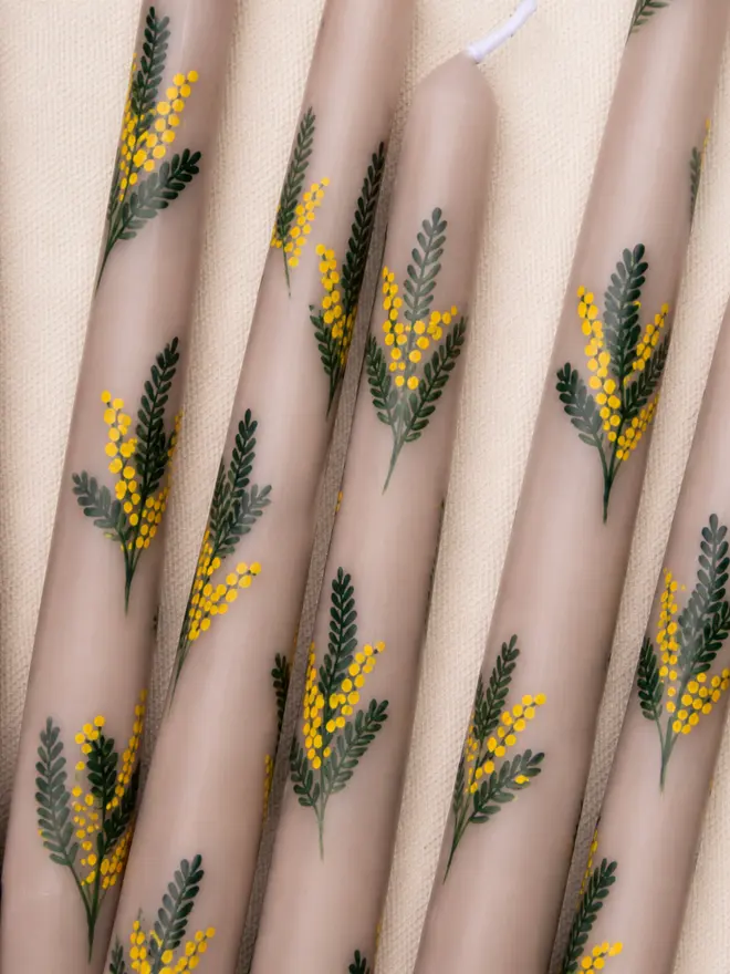 Close-up flat lay of hand-painted oat-coloured taper candles decorated with delicate mimosa flowers and green leaves on natural linen fabric.
