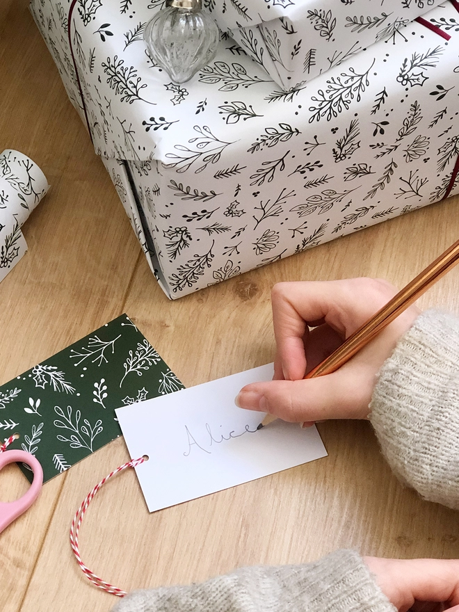 A gift wrapped in white wrapping paper with a Christmas botanical design lays on a wooden floor while a gift tag is being written beside it.