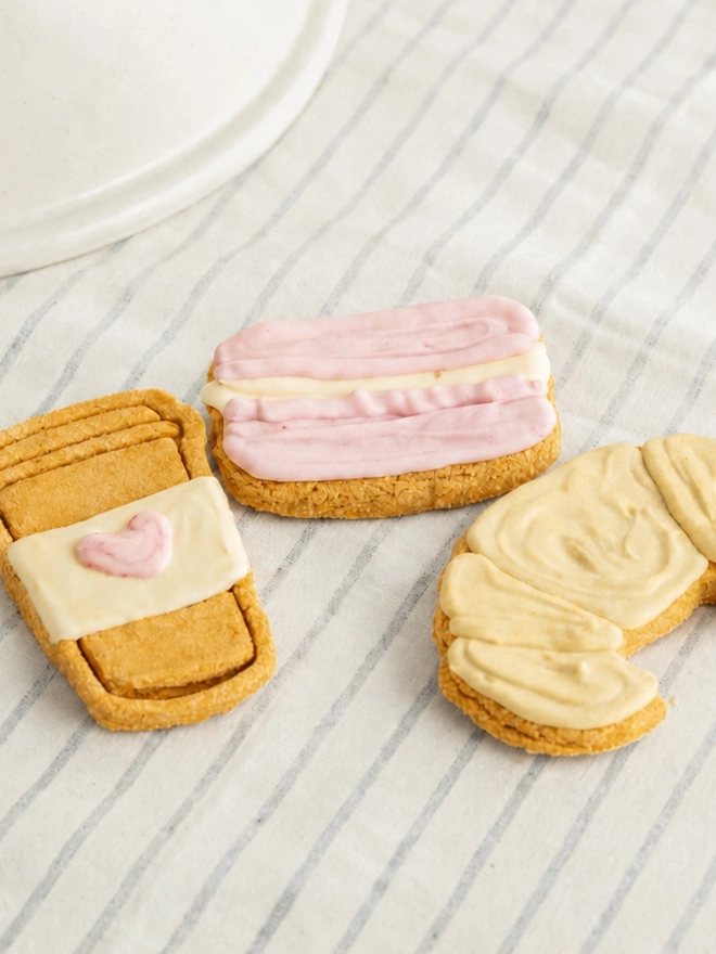 Parisian Bakery iced dog biscuit trio styled on striped fabric beside a ceramic plate.