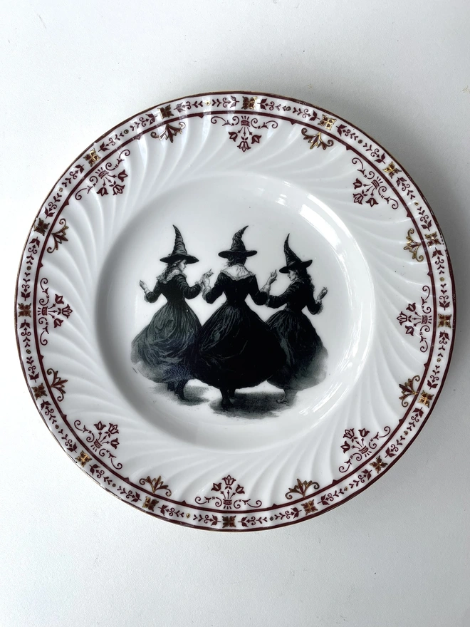 a coven of 3 witches dance hand in hand, the image is victorian in black and white and is printed directly onto an ornate vintage plate