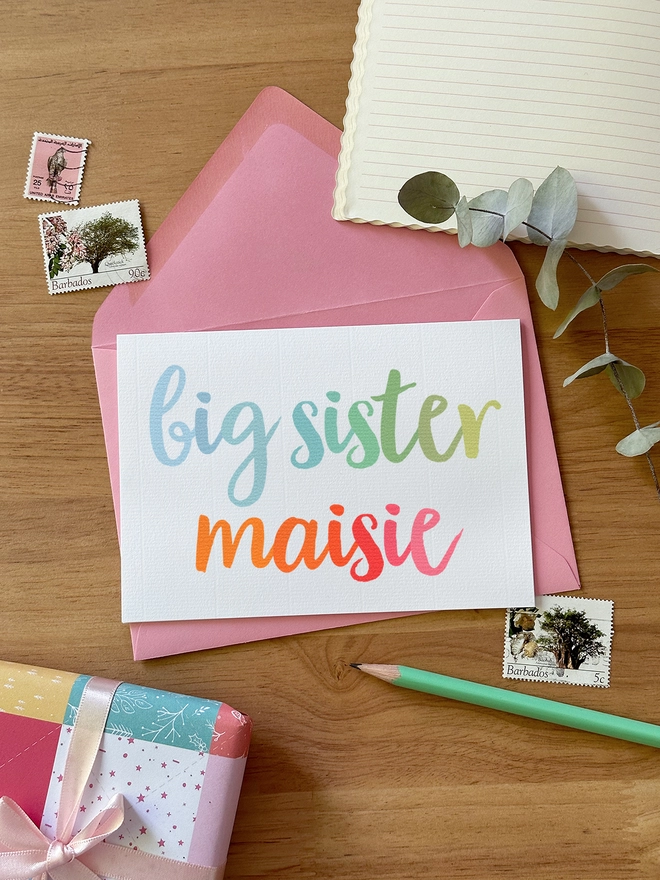 A personalised new baby sibling card with 'big sister' wording in pastel rainbow colours lays on a green envelope on a wooden desk.