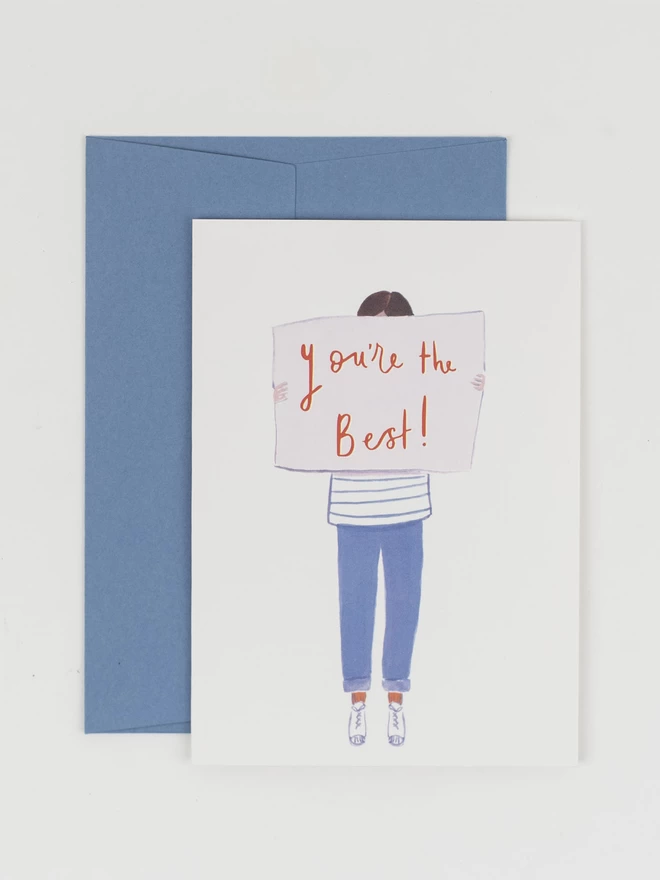 A greeting card featuring an illustrated person holding up a sign that covers their face. The sign reads "You're the Best". 