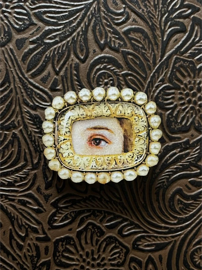 lovers eye brooch, the eye portrait is surrounded by a gold and pearl border, printed straight onto wood, with brass clip pin