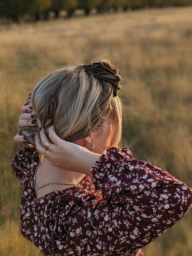 A caper green velvet fabric wrap headband. It is modelled on a blonde woman looking into the sunset in a field as she adjusts her hair.