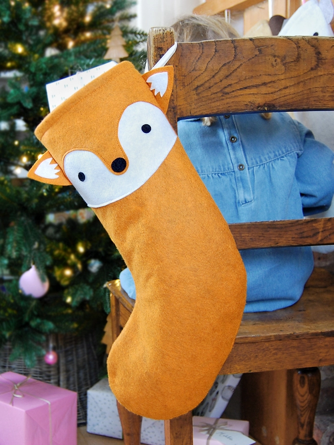 A handmade felt fox stocking hangs on a wooden chair, where a little girl sits, in front of a Christmas Tree.