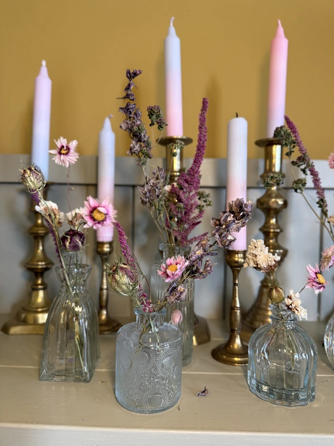 a selection of dried flowers in glass bud vases on a shelf with candles in brass candlesticks